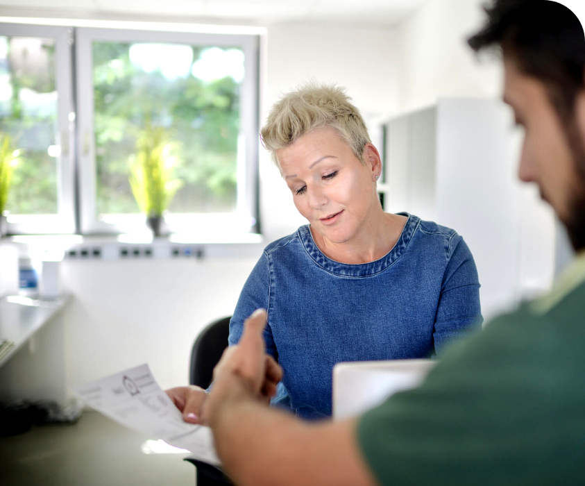 Frau mit blonden, kurzen Haaren, die in einem modernen Büro sitzt und einem Mann gegenüber sitzt, während sie Dokumente bespricht.